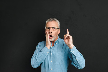 Image of excited retired man 60s with grey hair and beard in shirt shouting in surprise have idea and pointing index finger upward on copyspace, isolated over black background