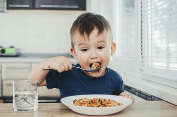 beautiful cute baby eats rice with a spoon in the kitchen, very fun
