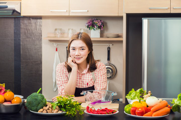 Proud attractive asian female cook standing in her home kitchen and smiling at camera, with various vegetables and fruits on the counter top