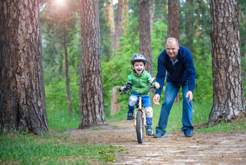 Little kid boy of 3 years and his father in autumn forest with a bicycle. Dad teaching his son. Man happy about success. Child with helmet. Safety, sports, leisure with kids concept.