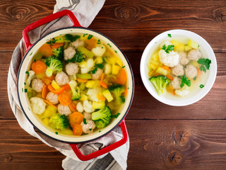 large metal cast red pan and plate with delicious hot soup of vegetables and meatballs, cauliflower, broccoli, carrots, peppers, potatoes, healthy diet food. Top view, wooden background