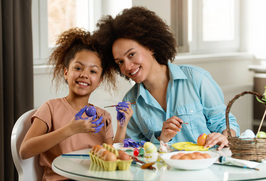 Beautiful African American woman and her daughter coloring Easter eggs at the table  