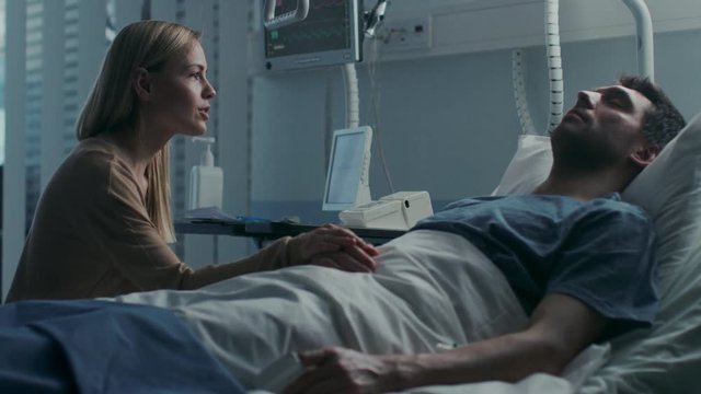 In the Hospital, Worrying Wife Sits Beside the Bed where Her Sick Husband Lies. She Holds His Hand, Talks to Him and Hopes for Recovery. Shot on RED EPIC-W 8K Helium Cinema Camera.