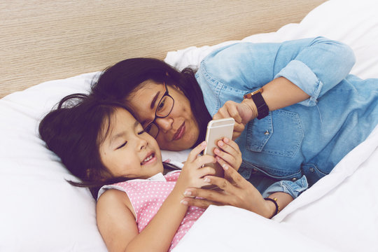 young mom and her little daughter are using a smartphone on bed at home