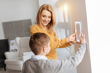 Latest technology. Delighted positive nice woman smiling and looking at her son while pointing at the control panel in the smart house