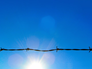 Wire fencing against a blue sky and sun.
Black horizontal line of sharp-cut silhouette and bright sunlight.