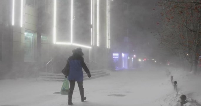 A woman is going through the blizzard in the street in a winter night