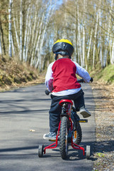 Little child boy cycling on bicycle in green park outdoor in spring. A child is riding a children's bike with support training wheels wearing safety helmet
