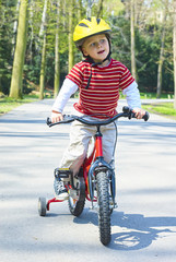 Little child boy cycling on bicycle in green park outdoor in spring. A child is riding a children's bike with support training wheels wearing safety helmet
