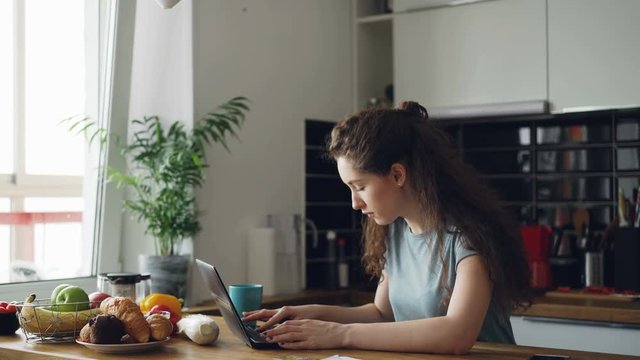 Young concentrated cheerful woman working in kitchen typing laptop computer during breakfast in the morning