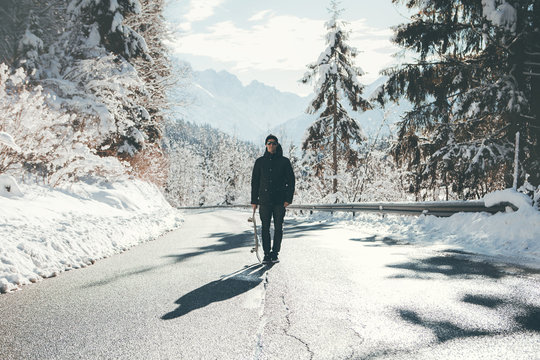 Skateboarder standing in the middle of the icy road  between the mountains, surrounded by the snow