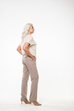 Full length portrait of beautiful adult woman 70s with gray hair and casual clothing looking aside with arms in pockets, isolated over white background