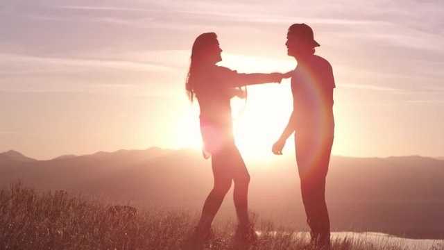Man stading on mountain top as woman walks over to hug him during colorful sunset lighting up the landscape.