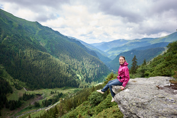 Happy beautiful woman tourist sitting on rock edge, looking to the camera, enjoying breathtaking view of green grassy slopes and mountains with trees, fir trees and pines in Romania