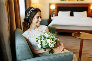 Elegant charming young brunette bride in the hotel room  is sitting on sofa in a wedding dress with a big bouquet of flowers, daydreaming, in the interior Waiting for groom. detailed retouch