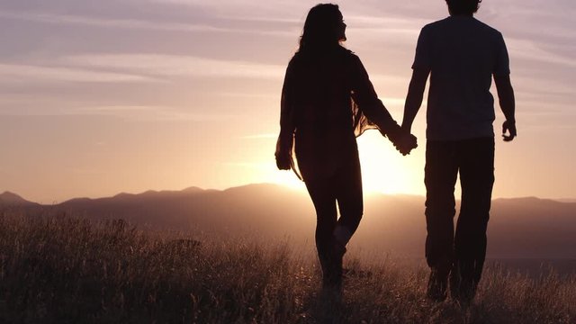 Slow motion of couple walking on hill top towards colorful sunset as they turn and kiss each other.