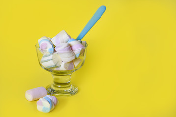 Sweet marshmallows in a glass with a blue spoon in a yellow background