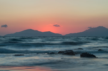 Obraz premium A long exposure of the sea at golden hour, as the sun sets behind the mountains. The sea looks ethereal as it breaks over the rocks and beach.