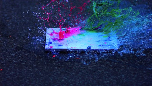 Slow Motion footage of a water-balloon hitting a canvas board with red green and blue paint on it causing spectacular splatters