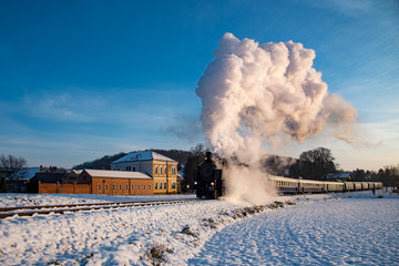 A steam engine in Austria as it steams up a hill near a village on a clear day with blue sky and the ground covered in snow.