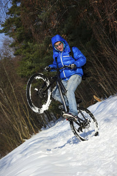Extreme cyclist standing on rear wheel of his mountain bike, winter sport, cross country biking on snowy road near forest in cool sunny day 