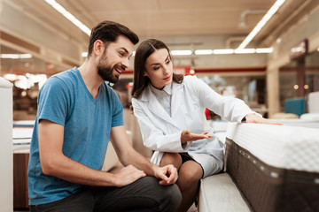 Naklejka premium Lovely woman consultant demonstrates orthopedic mattress to confident man in furniture store.