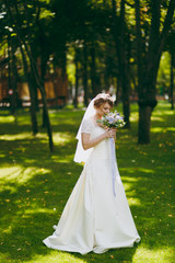 Beautiful wedding photosession. Elegant young bride in white dress and veil with beautiful hairdress with bouquet of flowers and ribbons near trees on wedding walk in the big green park on sunny day