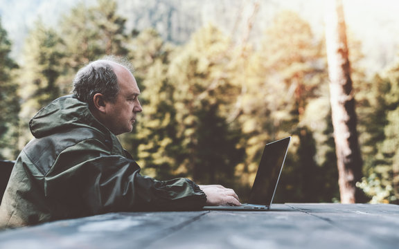 Side view of an adult male forester with a bald patch and grey hair, in the coverall, working on his laptop while sitting at a wooden table outdoors with forest in a defocused background