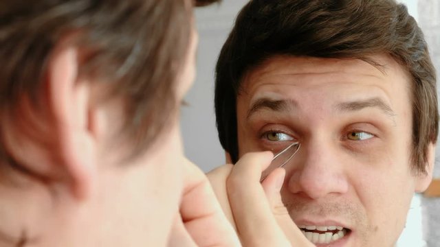 Young man plucking his eyebrows with tweezers and and winces from the pain. Styling eyebrows.