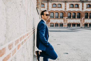 Fototapeta na wymiar .Young elegant man leaning on a brick wall, dressed elegant to work in a dark blue suit on a sunny day. Businessman. Lifestyle.