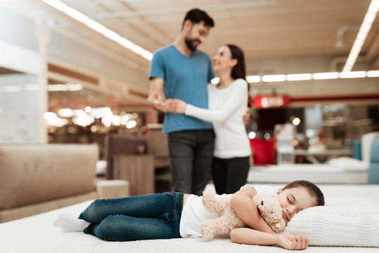 Young Happy Couple In Background Enjoying Sleeping Little Girl. Choosing Mattress In Store.