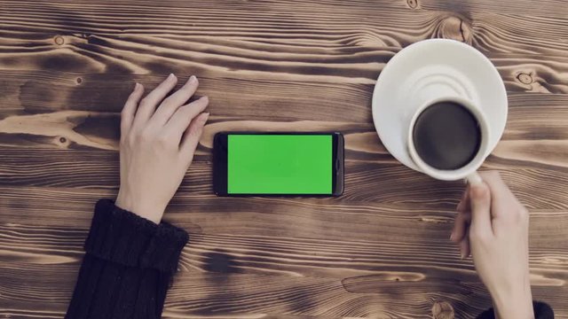 White cup coffee and smartphone with green screen in horizontal position on wooden table. Hands of young girl in pullover. Women drinks coffee, looks and scrolls finger of menu mobile app