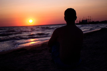 silhouette of a man who sits on the sand near the sea, in the rays of the setting sun near the sea. place under the text, relax on the sea
