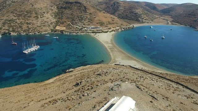 Flight over white church of St. Luke and the mountain with arid bushes towards yellow sand two-way Kolona beach with anchored yachts at hot bright summer day. Kolona Beach, Kithnos island, Greece