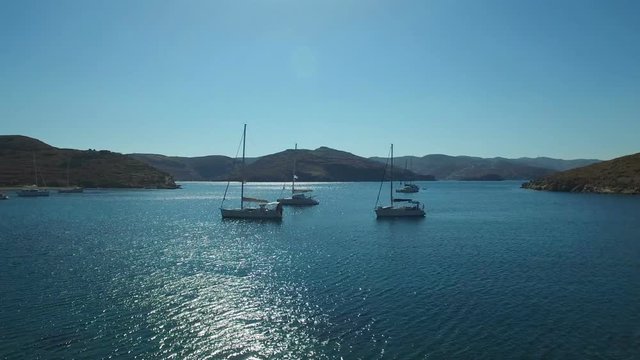 Luxury yachts are anchored in the lagoon of the Mediterranean Sea near the Kolona beach of Kitnos island. Apokrisi, Cyclades, Greece. 4k Aerial footage.