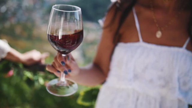 Hand of young unrecognizable female in white dress shakes glass of red wine outdoors on hot summer sunny day
