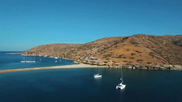 Flying above luxury yachts anchored in meditarranean sea lagoon near the Kolona beach of Kithnos island towards mountain with brown stones and arid bushes at hot sunny day with clear blue sky. Greece