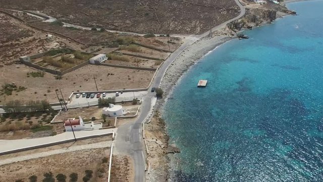 Car moves along Aegean sea coastal road among dusty brown stones, arid bushes. Landscape of Greek Cyclades and Aegean sea. Faros Armenistis, Mykonos Island, Greece. Aerial.
