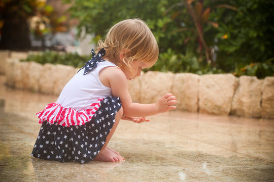 Cute Adorable Two Years Old Toddler Girl with Blonde Hair and Big Blue Eyes in Summer Dress with American Flag Print  is Dancing at the Rain, by the Pool Area in Cancun Resort in Mexico, March 2018