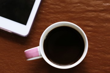 Top view of black coffee and part of tablet body closeup on rough brown texture background