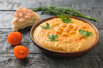 Clay bowl with fresh hummus, a piece of pita bread and sliced tomatoes on a wooden table.