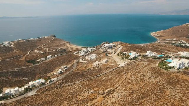 Flying down the hill with luxury white villas, swimming pools surrounded by brown stones, boulders, arid bushes, dusty roads with cars towards Aegean sea coastline. Cyclades. Faros Armenistis, Mykonos