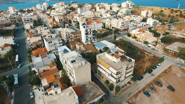 Flying over buildings of small town in southern Europe at sunset. Low-rise buildings, flat roofs, narrow streets, small cars, public squares, gardens. Aegean sea. Lavrio town, Attica, Greece. Aerial.