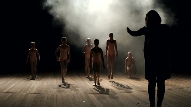 Woman choreographer teaches young children dance and ballet on stage in the dark on a black background. Classical school of ballet.