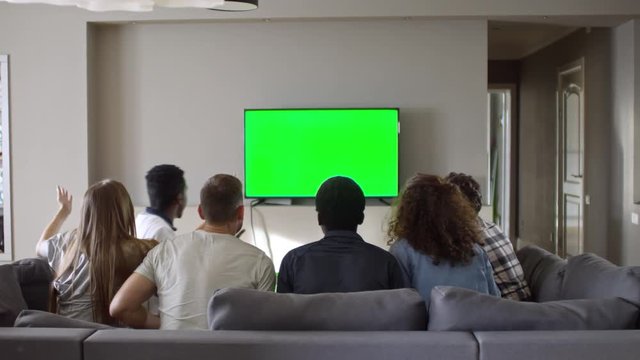 Rear view of young male and female soccer fans sitting on couch and watching sport match on chroma key TV screen. Friends standing up, raising hands and embracing while celebrating victory