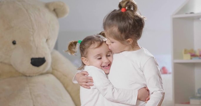 Portrait of two sisters in their bedroom smiling and kissing, in the background the stuffed animals and the dollhouse. Concept of: family, sisters, home and happiness.