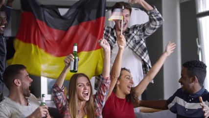 Group of multi-ethnic friends sitting together on couch with flag of Germany, drinking beer, embracing and yelling while watching sport competition on TV and celebrating goal