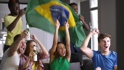 Company of excited young sport fans sitting together on couch with flag of Brazil, drinking beer, embracing, yelling and giving high-five while watching match on TV and celebrating goal