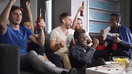 Multi-ethnic group of young men and women watching sport match on TV at home, raising arms, yelling, toasting with beer and celebrating goal