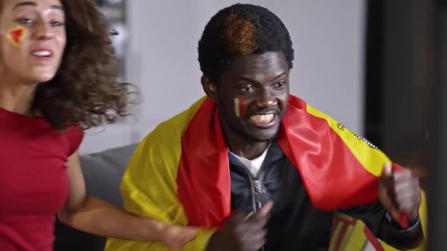 Excited African man holding flag of Spain, yelling and embracing his girlfriend while watching soccer on TV at home